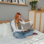 Mulher utiliza um laptop sentada na cama, em um quarto residencial.