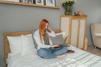 Mulher utiliza um laptop sentada na cama, em um quarto residencial.
