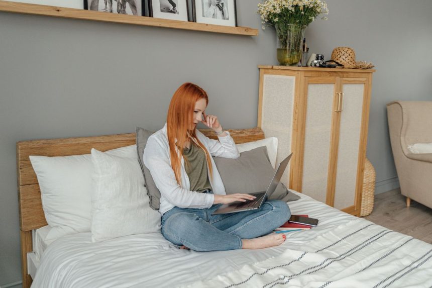 Mulher utiliza um laptop sentada na cama, em um quarto residencial.