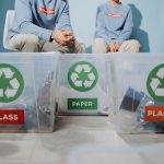 Pessoas organizam materiais recicláveis em recipientes separados para vidro, papel e plástico.