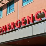 Entrada de um pronto-socorro hospitalar moderno, com acesso coberto e identificação em letras vermelhas.